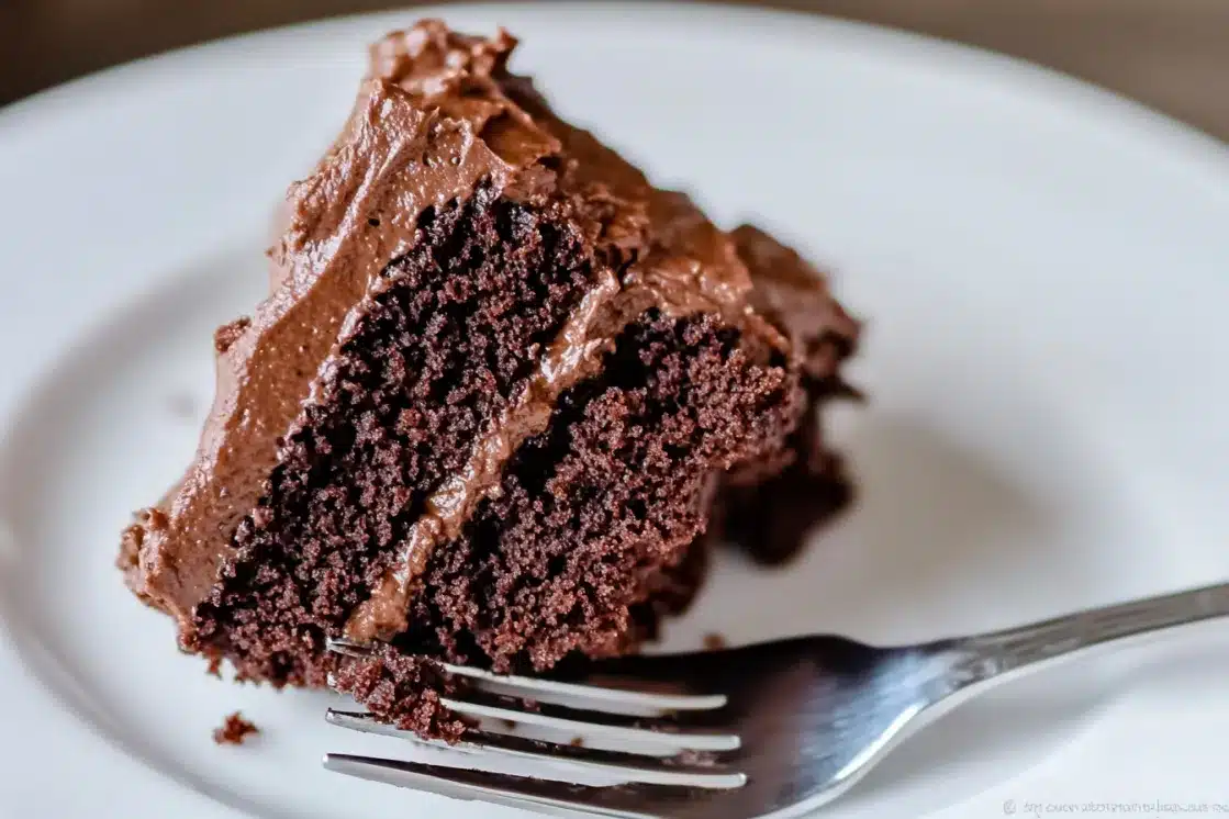 Moist chocolate cake slice with rich chocolate frosting on white plate, showing tender crumb and fudgy texture