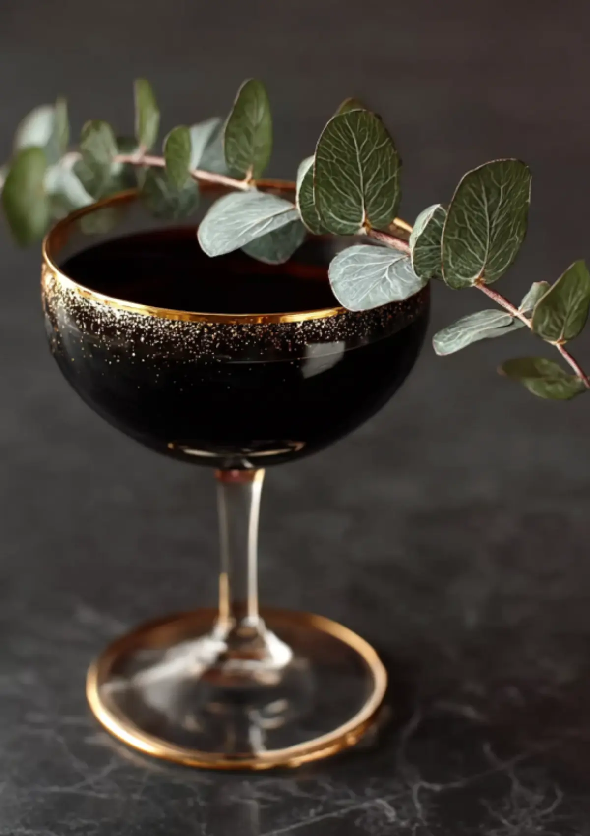 Black cocktail in gold-rimmed coupe glass with multiple eucalyptus sprigs as garnish