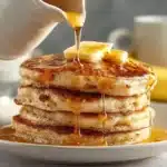 Stack of fluffy golden banana pancakes on white plate with butter on top and maple syrup being poured from white pitcher, banana visible in background