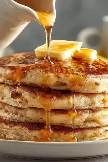Stack of fluffy golden banana pancakes on white plate with butter on top and maple syrup being poured from white pitcher, banana visible in background