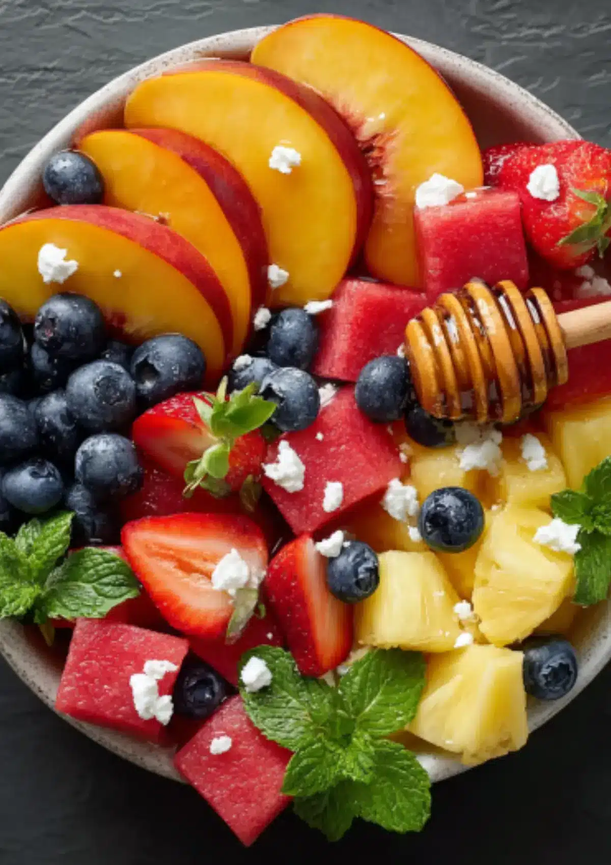 Colorful fruit salad bowl with peaches, watermelon, strawberries, blueberries, pineapple, feta cheese, mint leaves, and honey drizzleColorful fruit salad bowl with peaches, watermelon, strawberries, blueberries, pineapple, feta cheese, mint leaves, and honey drizzle