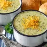 Two white enamel mugs filled with creamy zucchini soup topped with shredded cheddar cheese and fresh herbs, served alongside crusty bread rolls on a blue striped kitchen towel