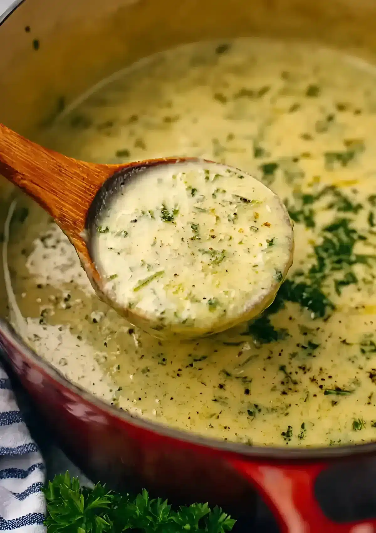 Wooden ladle holding a spoonful of creamy zucchini soup over a red Dutch oven pot, showing the smooth, herb-flecked texture of the finished soup