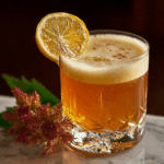 Detail view of layered amber cocktail with creamy foam top, burgundy flower and lemon garnish