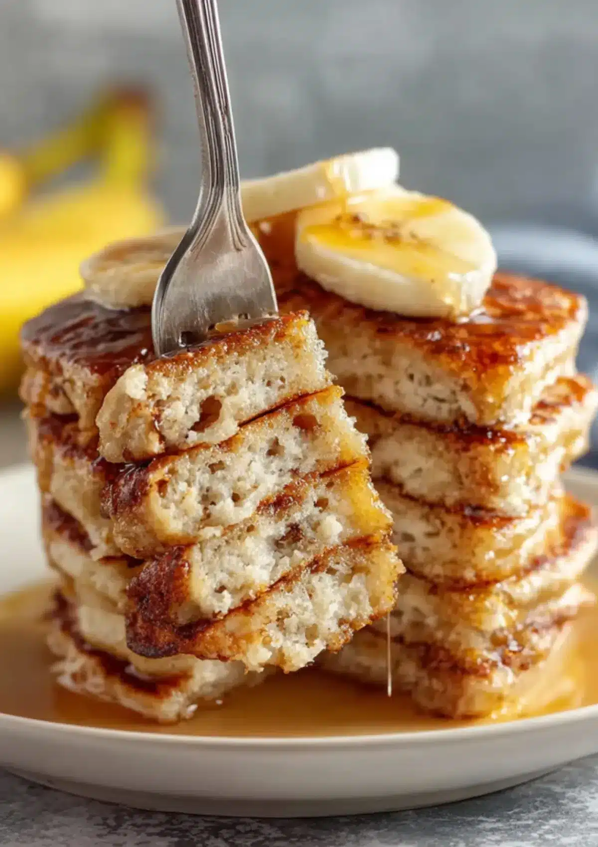 Silver fork holding bite-sized piece of banana pancakes showing internal fluffy texture with banana slices on top and maple syrup drizzling

