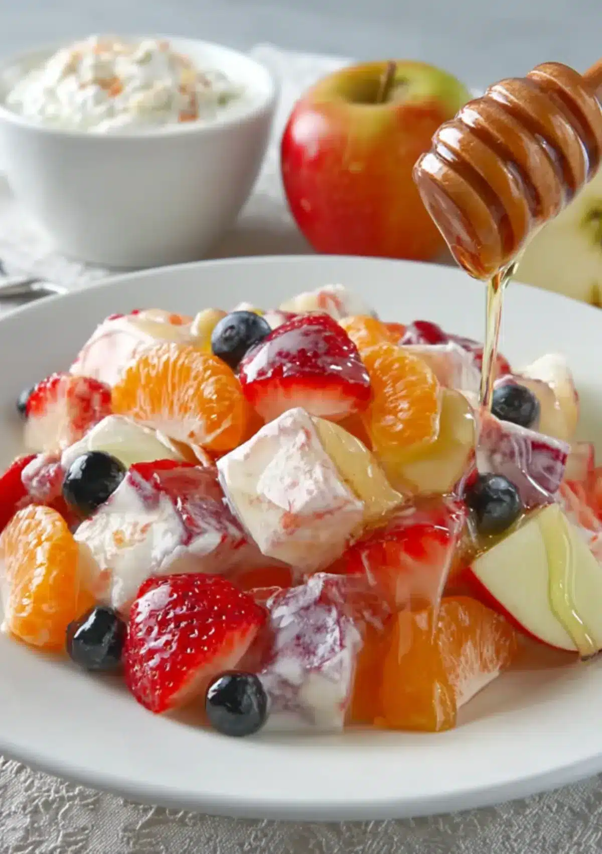 A plate of creamy yogurt fruit salad topped with strawberries, blueberries, apple slices, and mandarin orange segments, drizzled with golden honey from a honey dipper.