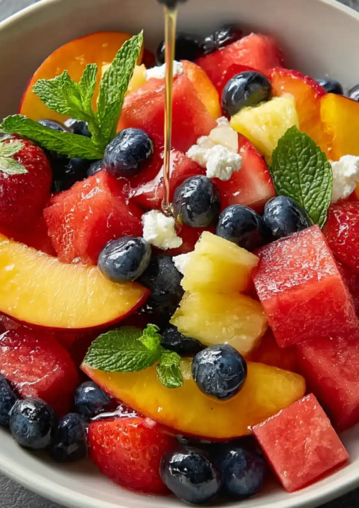 Fresh fruit salad with honey being drizzled over mixed berries, peaches, watermelon, pineapple, and mint
