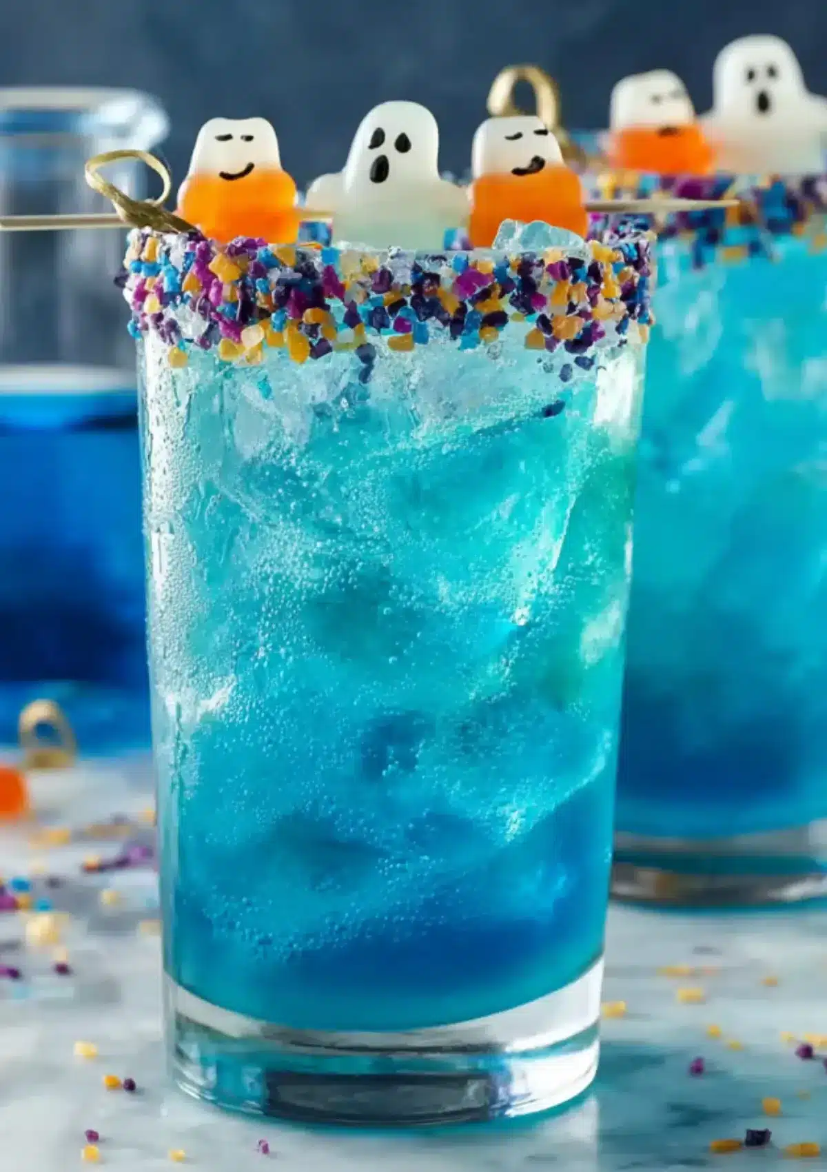 Tall highball glass filled with a bright blue drink and crushed ice, rim coated with colorful sprinkles, topped with gummy ghosts on a cocktail pick.
