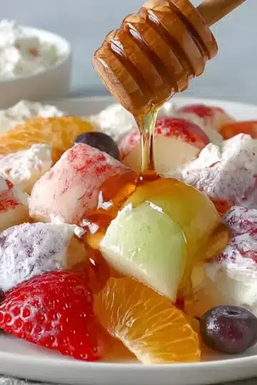Close-up of mixed fruit salad containing strawberries, orange segments, green grapes, blueberries, and white cheese cubes coated in colorful fruit juices, with honey being drizzled from a wooden honey dipper. A bowl of yogurt and an apple are visible in the background