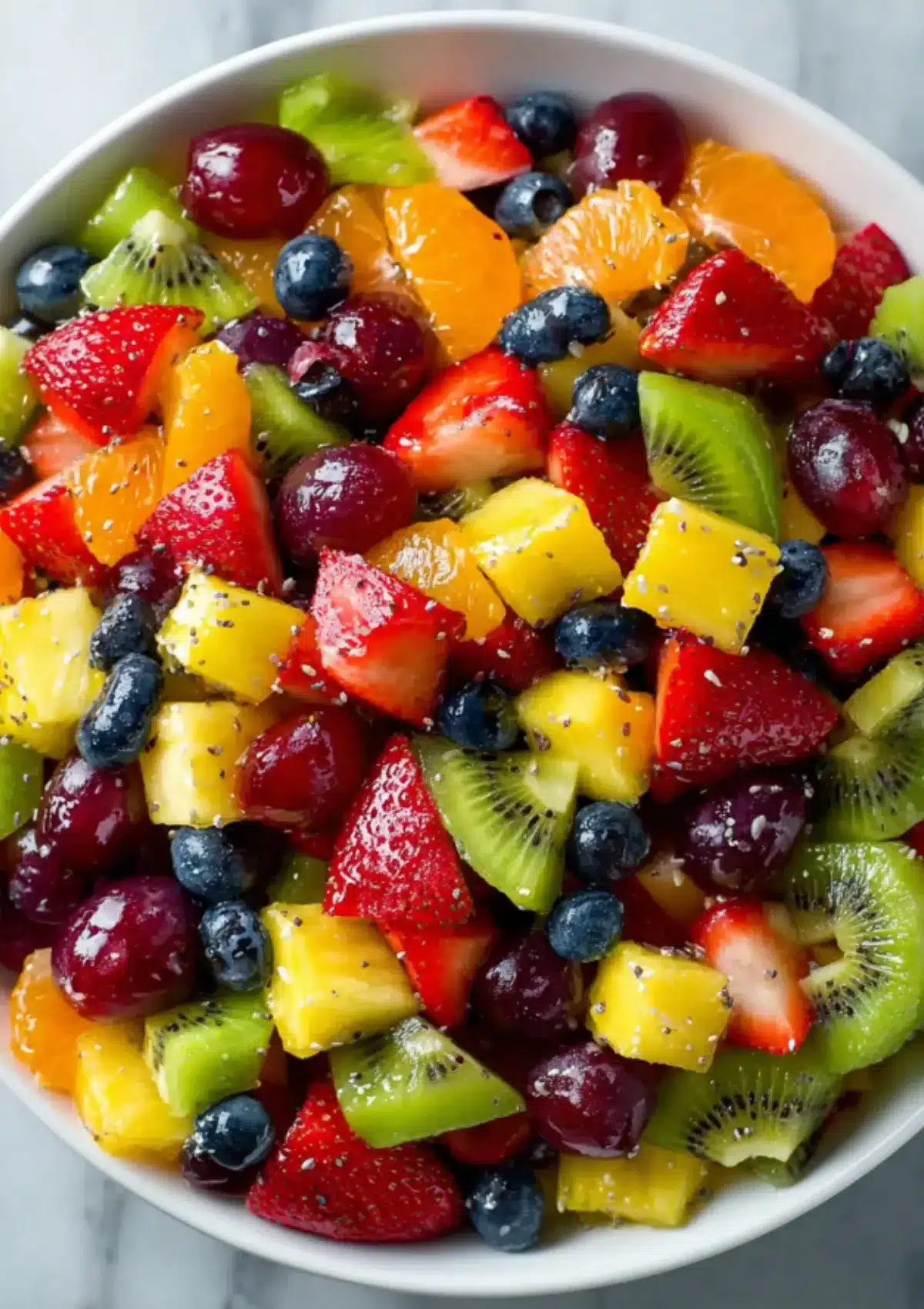 Close-up overhead view of vibrant fruit salad in white bowl showing cubed pineapple, strawberries, kiwi, blueberries, red grapes, and orange segments all glistening with honey lime dressing