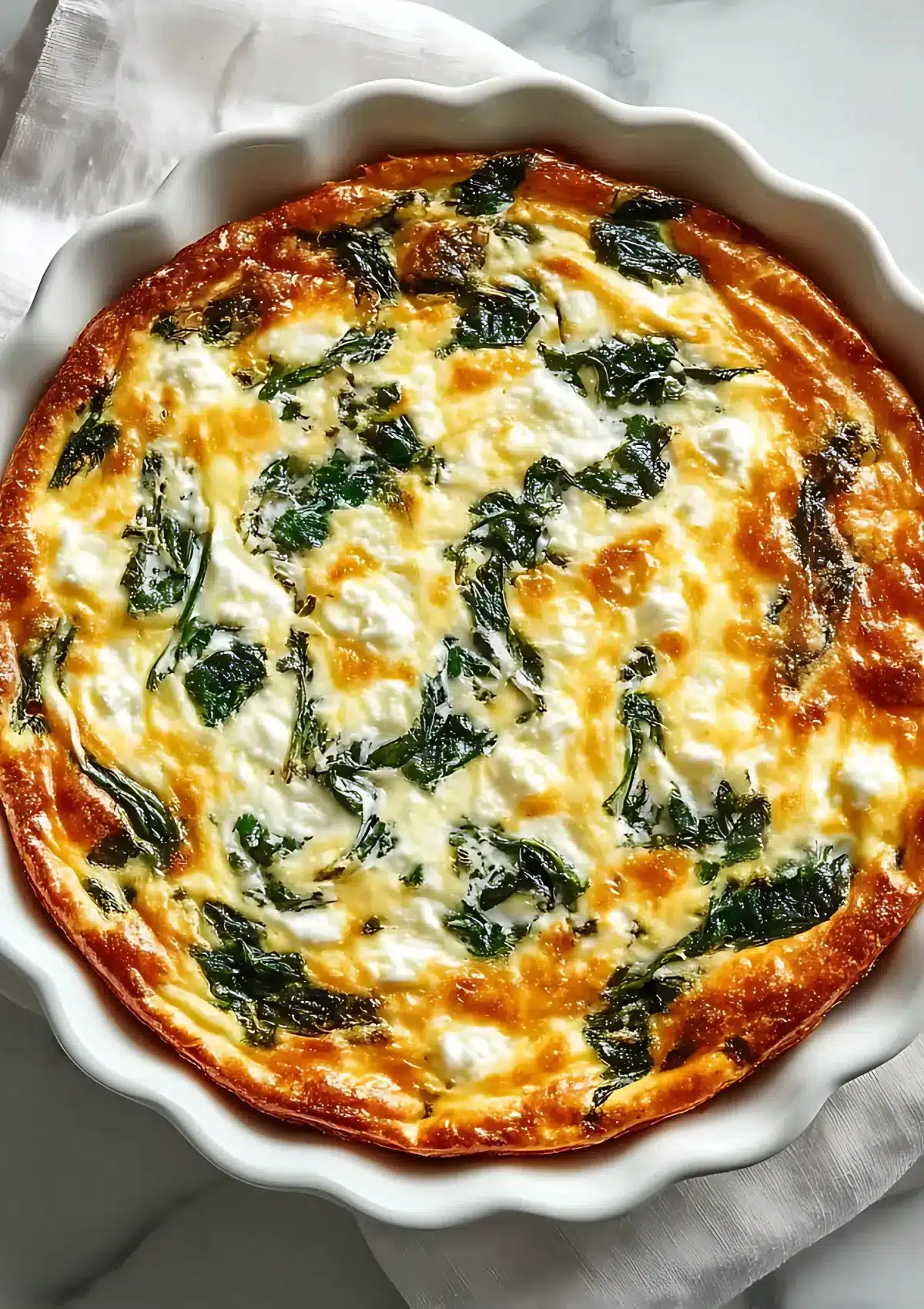 Freshly baked cottage cheese and spinach crustless quiche with a golden top and wilted spinach leaves, styled on white marble countertop.