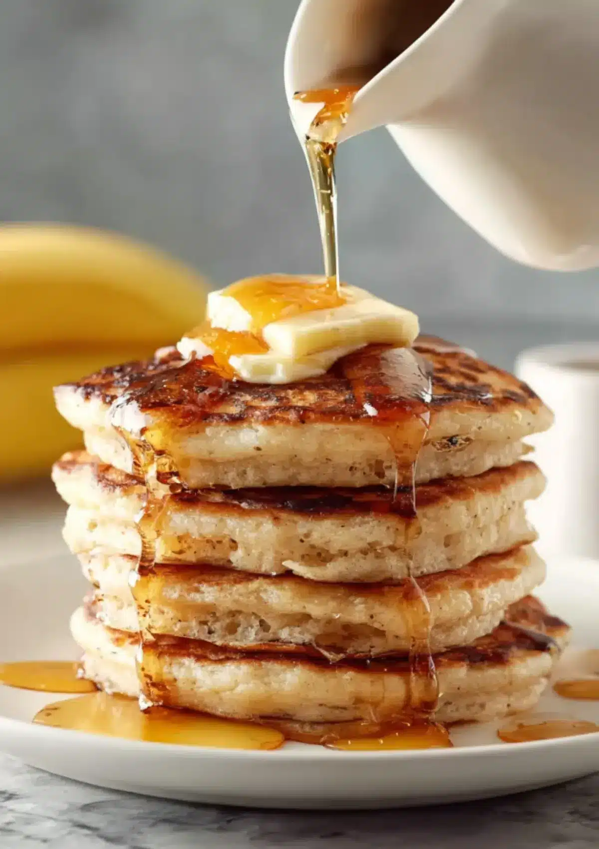 High stack of golden brown banana pancakes on white plate with square butter pat on top and maple syrup being poured from white pitcher, fresh banana in background


