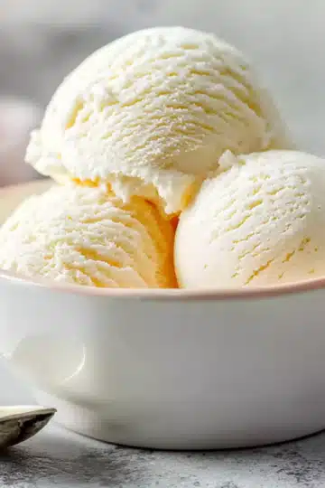 Three scoops of vanilla ice cream in a white ceramic bowl with a silver spoon beside it