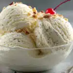 Vanilla ice cream scoops with nuts and vanilla bean specks in a clear glass bowl with cherry garnish