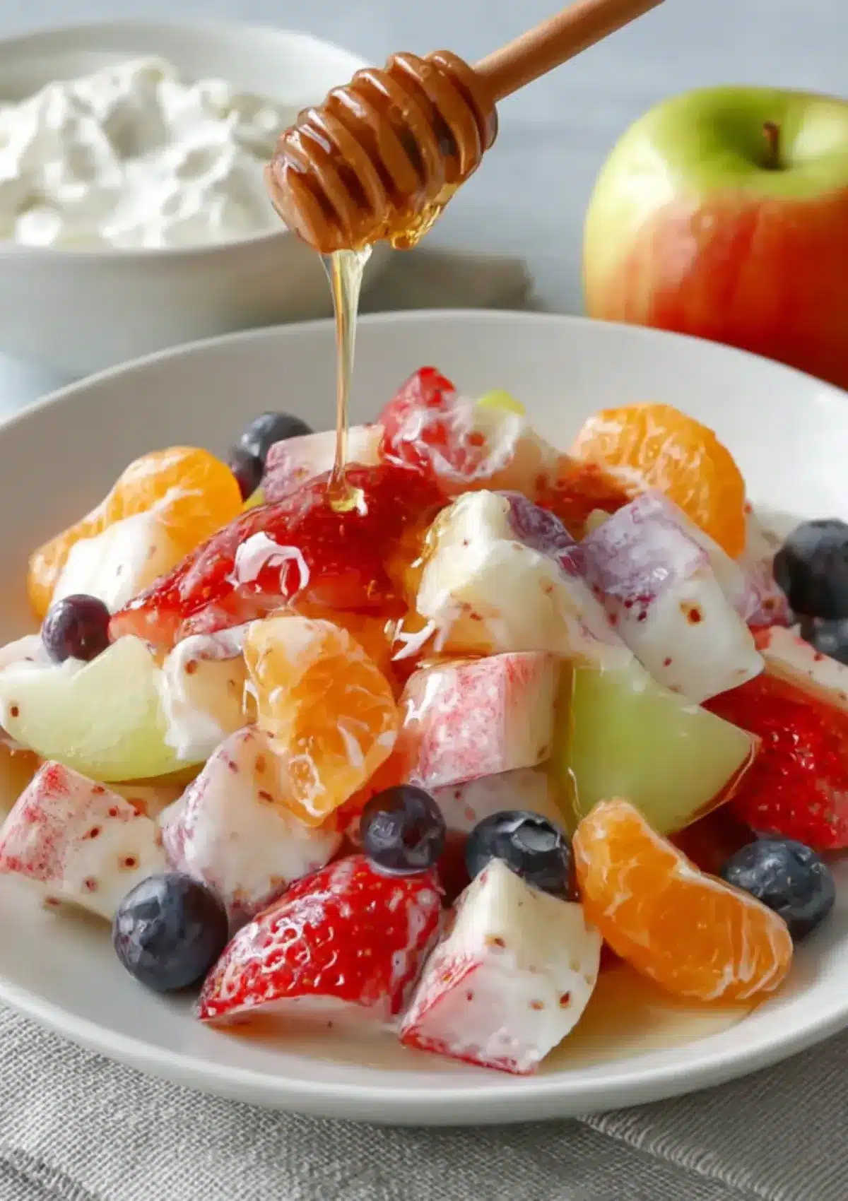  Close-up of a creamy yogurt fruit salad featuring apples, strawberries, blueberries, melon, and mandarin oranges, with honey being poured over the top.

