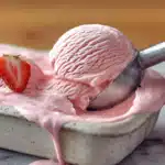 Ice cream scoop lifting pink strawberry ice cream from a white container with fresh strawberry slices as garnish