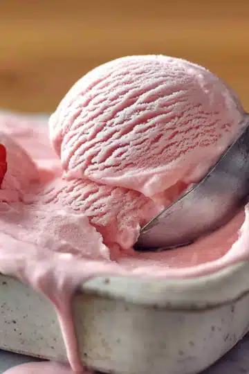 Ice cream scoop lifting pink strawberry ice cream from a white container with fresh strawberry slices as garnish