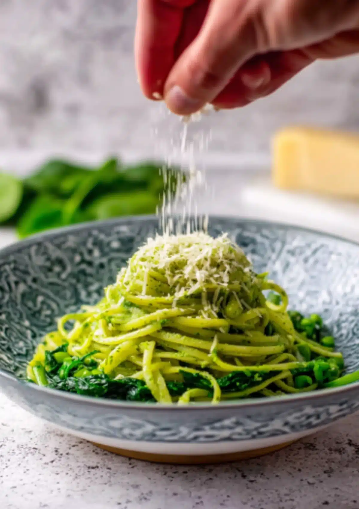 Hand sprinkling cheese over green pasta in decorative bowl