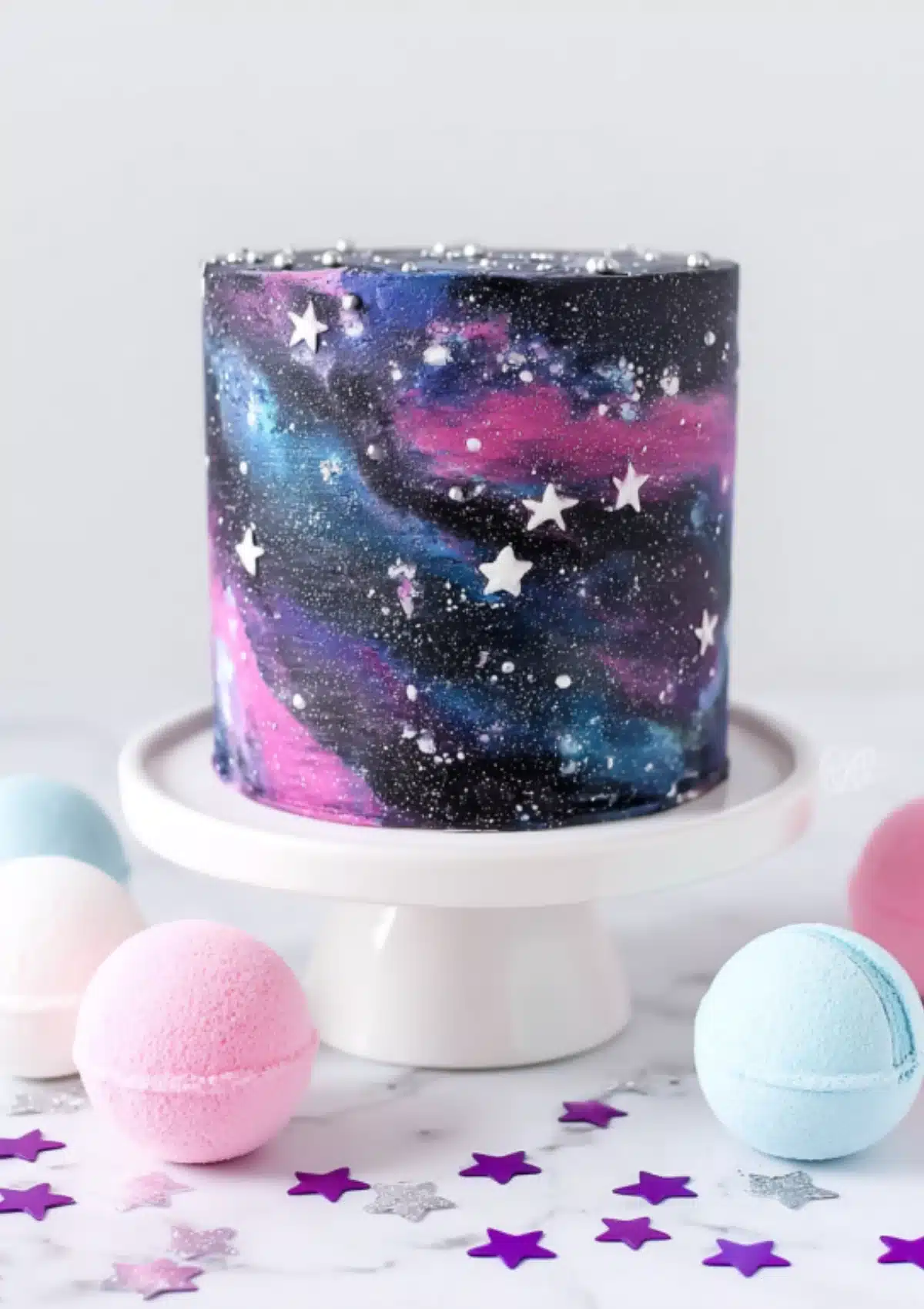 Galaxy cake on white stand with pink and blue bath bombs and purple star confetti