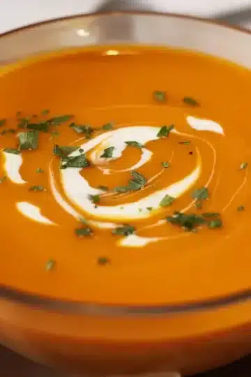 Hero shot of vibrant orange curried carrot soup with perfect cream swirl garnish and fresh herbs in rustic ceramic bowl with matching saucer, grilled flatbread on side