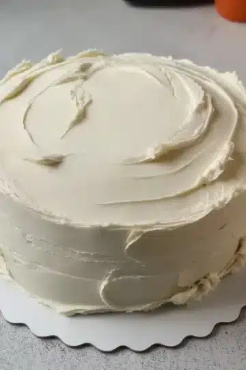 A small round cake covered in smooth, swirled buttercream icing on a scalloped white cake board over a gray countertop.