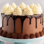 Chocolate layer cake with glossy dark chocolate drip and white cream swirls, placed on a blue cake stand with milk bottles in the background.