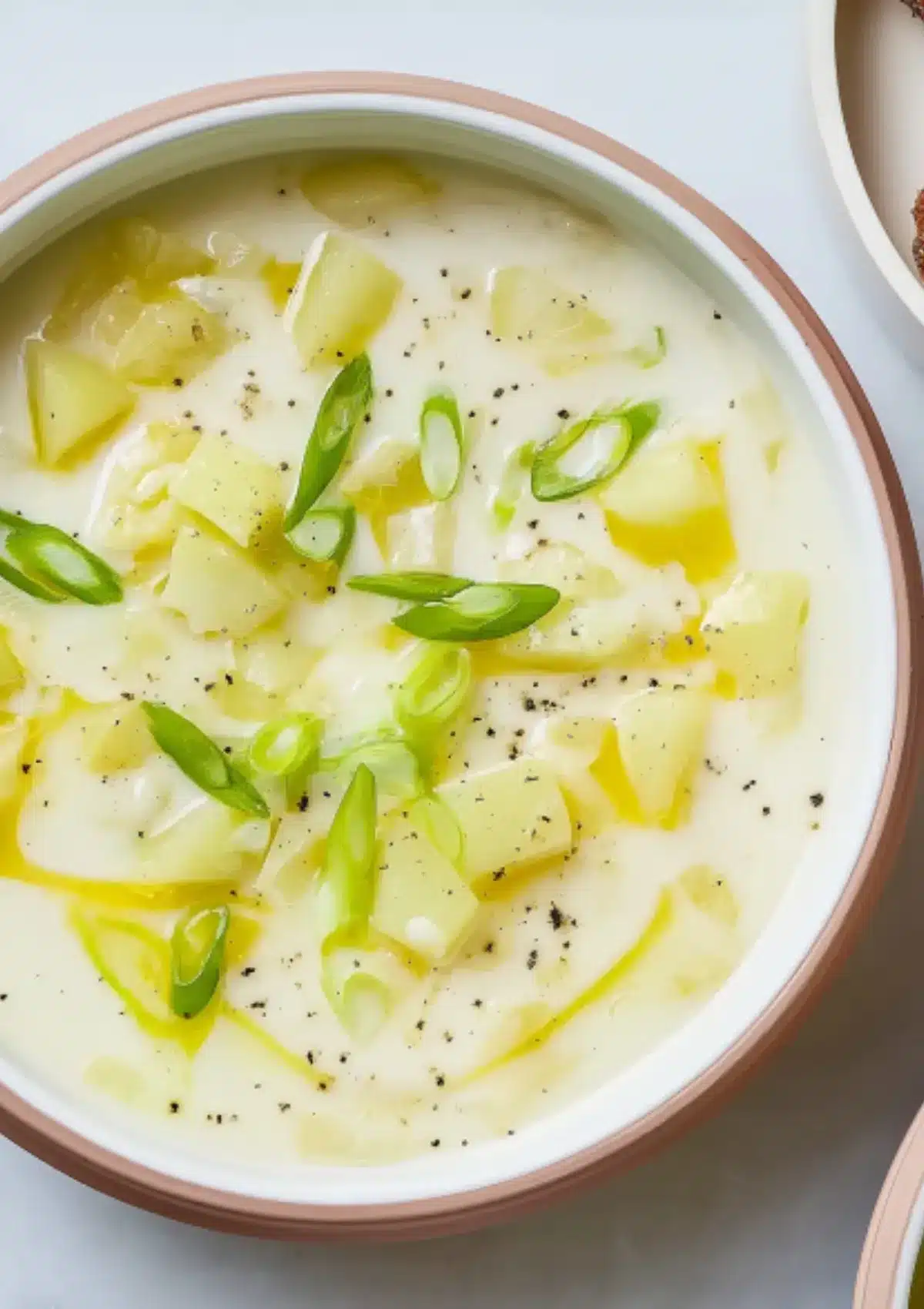 Top-down view of chunky potato leek soup in modern white bowl, featuring tender Yukon Gold potato pieces, vibrant green leek garnish, and olive oil finish on creamy ivory-colored soup base