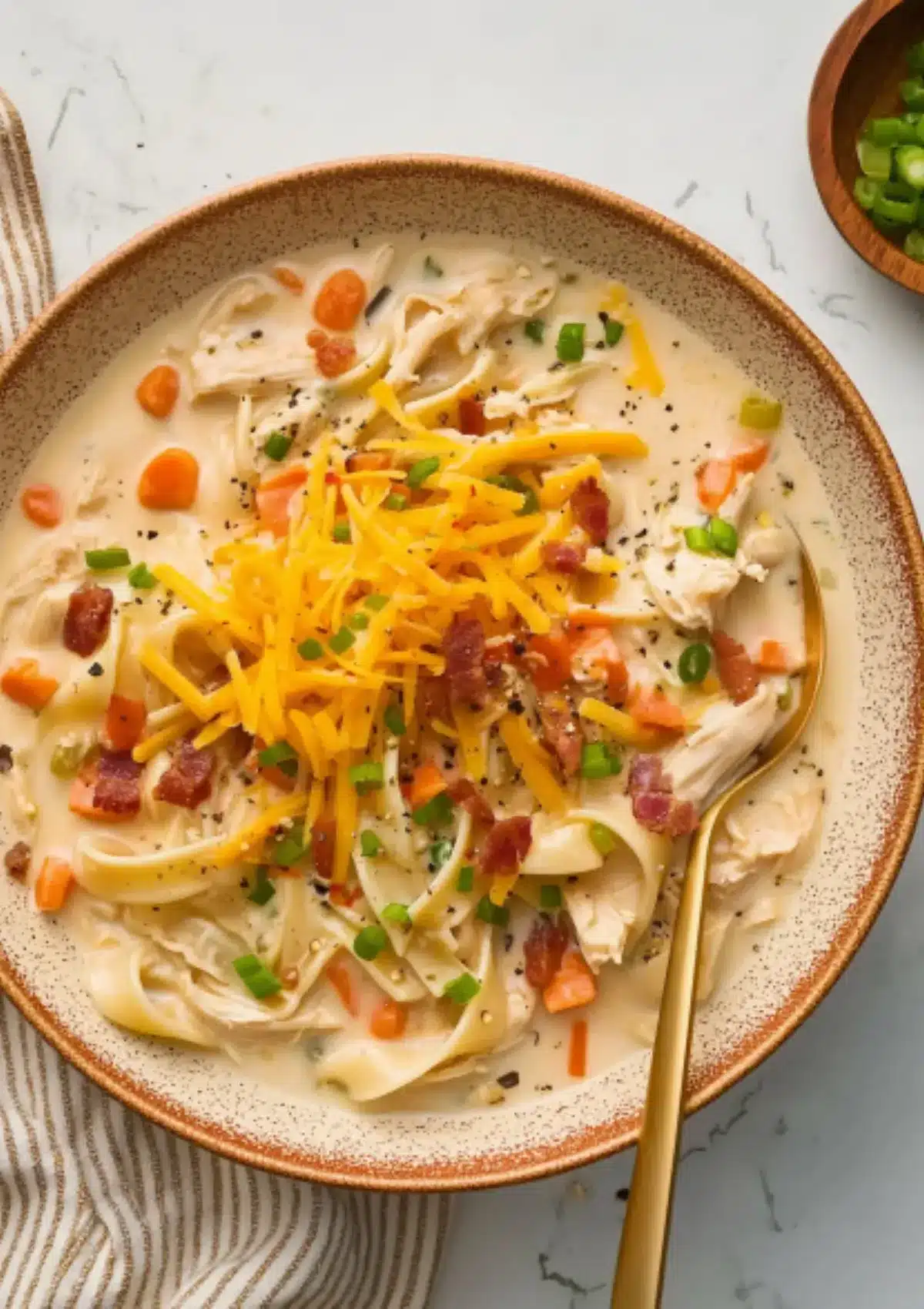 Crack chicken noodle soup served in speckled ceramic bowl with brown rim on white marble counter, garnished with shredded cheddar cheese, bacon crumbles, carrots, and green onions