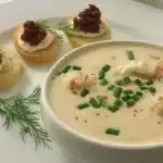 She-crab soup served in white bowl with rich cream broth, generous lump crabmeat pieces, fresh chopped chives, dill garnish, paprika seasoning, accompanied by three crostini with bacon jam and fresh dill on white plate
