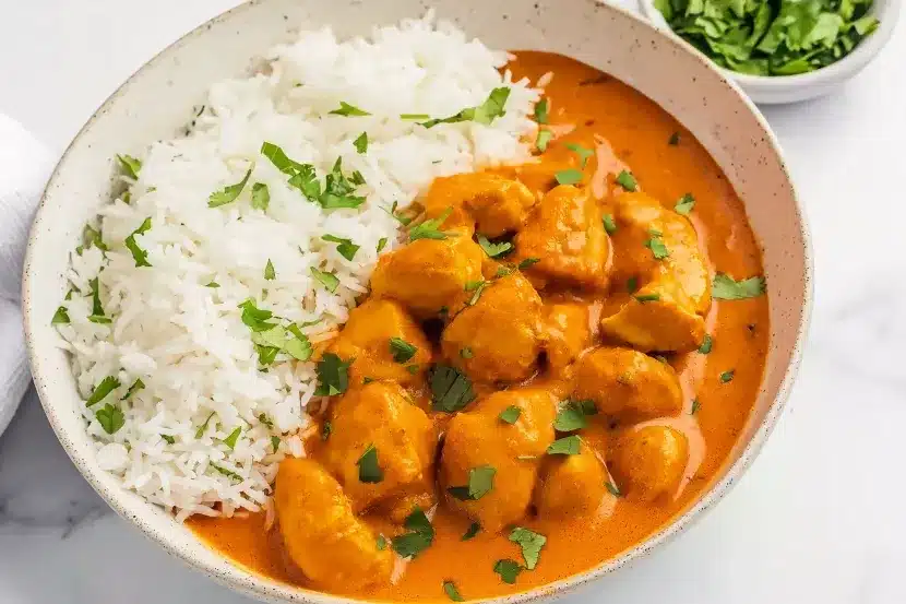 Overhead view of crockpot butter chicken with rice in speckled bowl on marble countertop - rich creamy curry with fresh cilantro garnish and herb bowl in corner