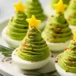 Christmas tree shaped deviled eggs with swirled green avocado filling, yellow bell pepper star toppers, and red pepper seasoning on white serving platter with rosemary