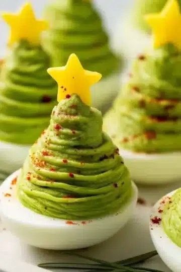 Christmas tree shaped deviled eggs with swirled green avocado filling, yellow bell pepper star toppers, and red pepper seasoning on white serving platter with rosemary