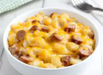 Slow cooker sausage and potato casserole in white bowl with golden diced potatoes, sliced sausage pieces, and melted cheddar cheese on rustic white wooden surface with green checkered napkin and fork
