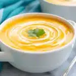 Professional food photography of sweet potato soup in white handled bowl with cream swirl design and cilantro leaf garnish, teal napkin and silver spoon beside bowl on white marble surface