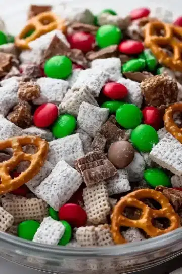 Glass bowl filled with reindeer chow mixture showing white powdered sugar coating on cereal pieces mixed with red and green M&M's, Reese's Minis, and mini pretzels