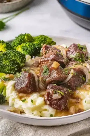 Plated serving of garlic butter crockpot steak bites with mashed potatoes and broccoli, showing a complete meal with visible slow cooker in background