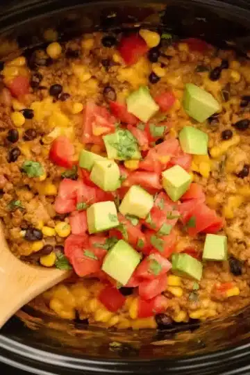 Oval black slow cooker containing Mexican taco casserole with ground beef, rice, black beans, yellow corn, melted cheese, garnished with fresh avocado and tomatoes, wooden spoon visible