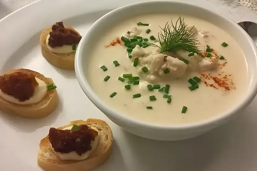 Elegant plating of South Carolina she-crab soup in white ceramic bowl with ivory colored cream base, lump crabmeat center, fresh dill sprig, scattered chives, paprika dusting, three bacon jam crostini on side