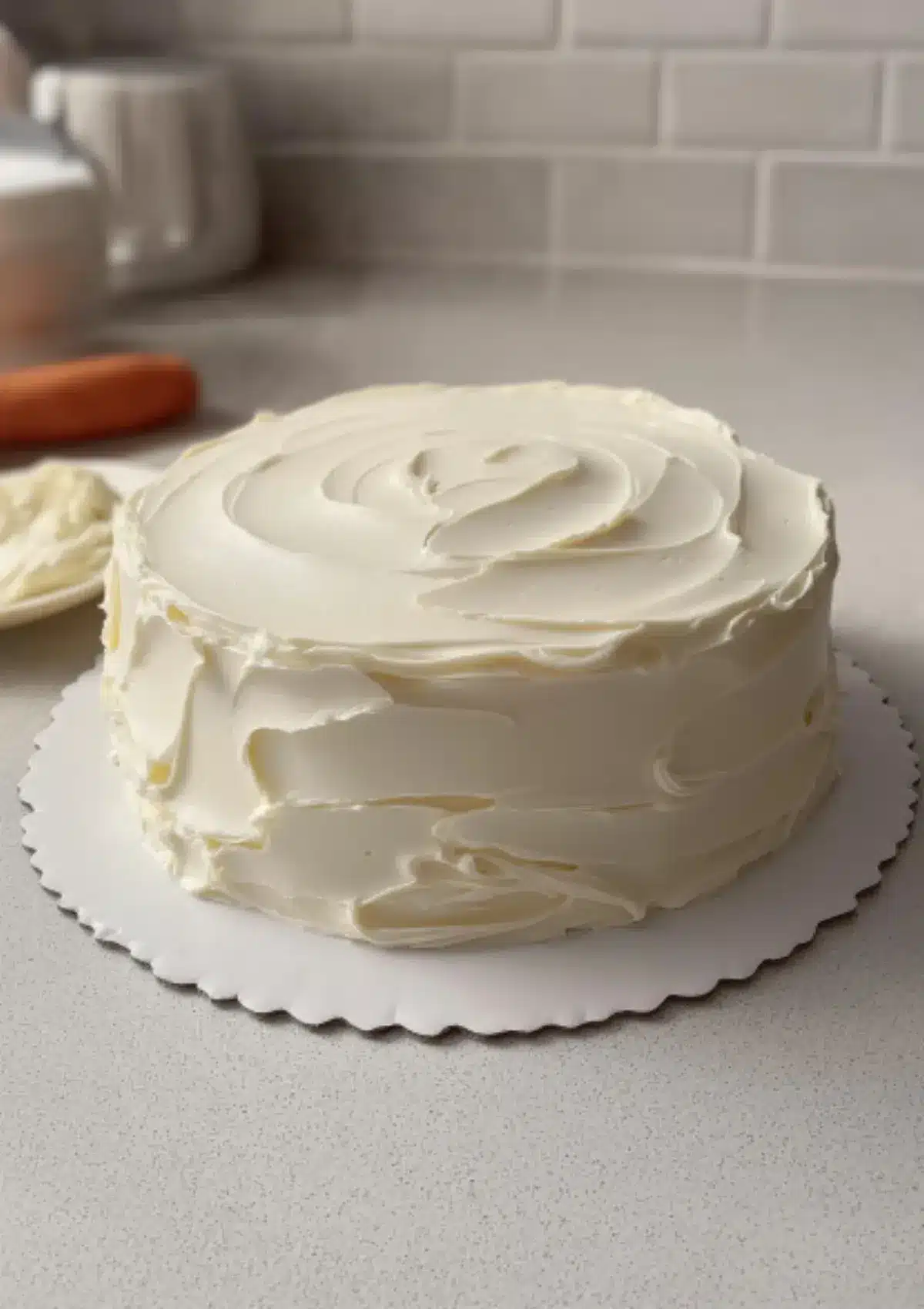 A vanilla layer cake covered in creamy buttercream icing, sitting on a white cake board on a kitchen countertop.
