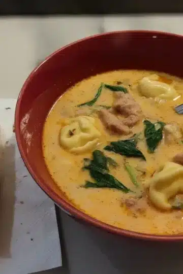 Individual serving of creamy sausage tortellini soup in burgundy bowl with garlic bread side, featuring tender tortellini and spinach in creamy base