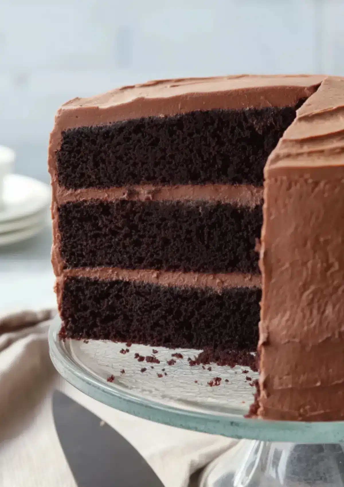 A close-up view of a sliced three-layer chocolate cake with rich dark layers and chocolate frosting on a clear glass cake stand.