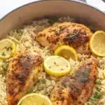 Bird's eye view of One Pot Lemon Herb Chicken and Rice showing three herb-crusted chicken breasts with Italian seasoning and black pepper, surrounded by fluffy white rice and fresh lemon slices in cream-colored enameled Dutch oven with gray handles