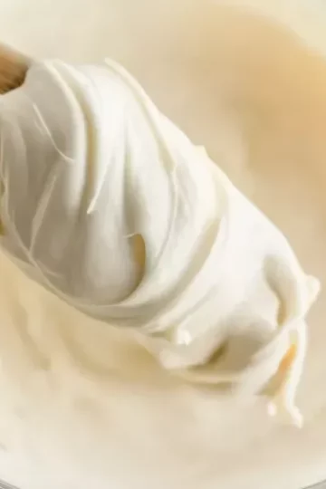 Wooden spoon holding a swirl of creamy vanilla buttercream frosting above a bowl.