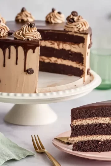 Full mocha cake with ganache drip and piped coffee buttercream on a white stand, paired with a plated slice and green glassware on a marble surface.
