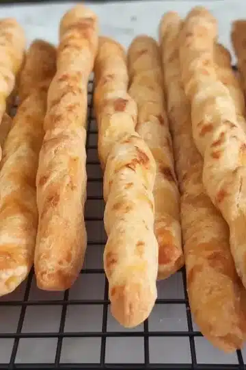 Diagonal view of golden brown cheddar cheese straws with speckled texture arranged on a black wire cooling rack
