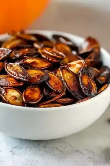 Air fryer pumpkin seeds with glossy caramelized coating in white bowl on white marble surface with orange pumpkin and scattered seeds