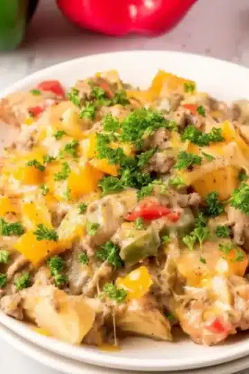 Generous serving of crock pot cheesesteak potato casserole on white plate topped with shredded cheddar cheese and fresh parsley with kitchen props in background