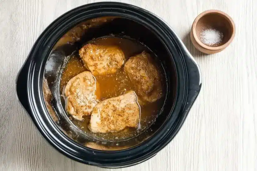 Overhead shot of four boneless chicken thighs cooking in black crock-pot slow cooker, surrounded by golden lemon-garlic sauce, showing caramelization and seasoning during cooking process on white wooden surfaceOverhead shot of four boneless chicken thighs cooking in black crock-pot slow cooker, surrounded by golden lemon-garlic sauce, showing caramelization and seasoning during cooking process on white wooden surface