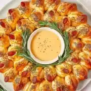 Beautifully styled pigs in a blanket Christmas wreath on white round plate featuring egg-washed crescent rolls wrapped around cocktail sausages in perfect circular wreath pattern, garnished with rosemary sprigs, everything bagel seasoning, and honey mustard dip in center bowl, placed on light marble countertop