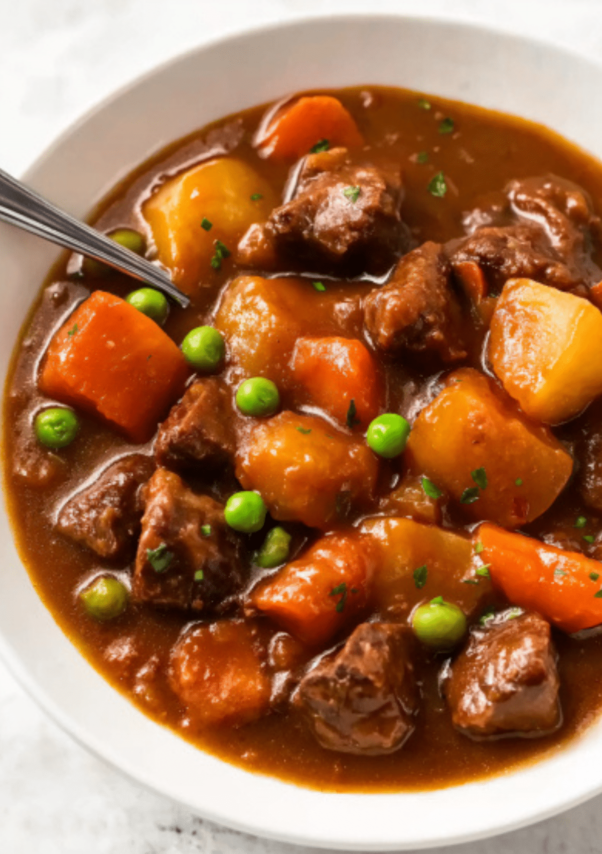 Appetizing close-up photograph of homemade slow cooker beef stew served in a white ceramic bowl. The stew features large chunks of tender braised beef, bright orange carrot rounds, golden yellow Yukon gold potato pieces, and vibrant green peas, all swimming in a rich, glossy brown gravy made with red wine and beef broth. Fresh parsley garnish visible. Silver spoon rests in the bowl. Shot against white marble background.